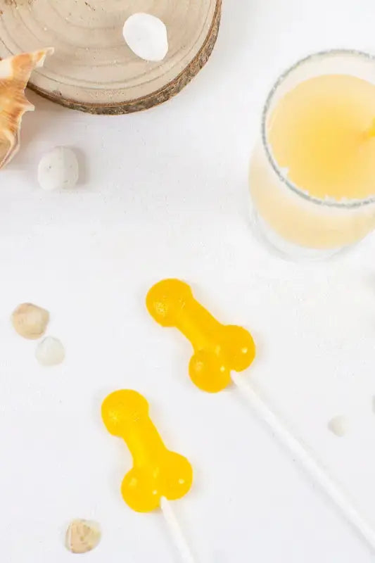 Sucette pénis alcoolisée goût pina colada sur une table avec un verre de boisson et des coquillages.
