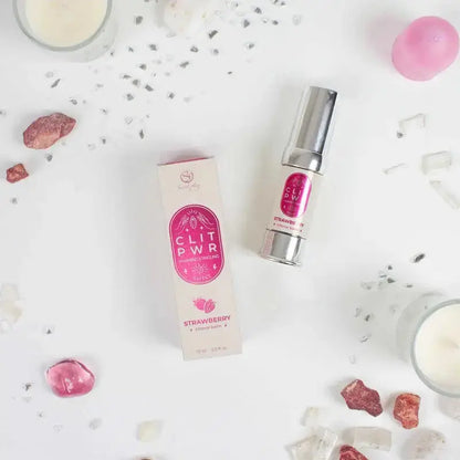 Baume clitoris Clit Pwr Fraise présenté avec son emballage sur une surface décorée de cristaux et de bougies.