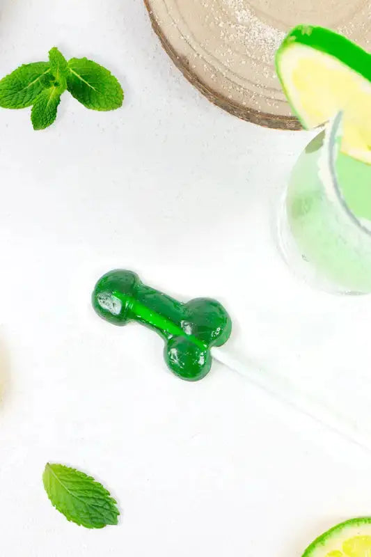 Sucette pénis goût mojito alcoolisée sur une table avec des feuilles de menthe et des quartiers de citron.